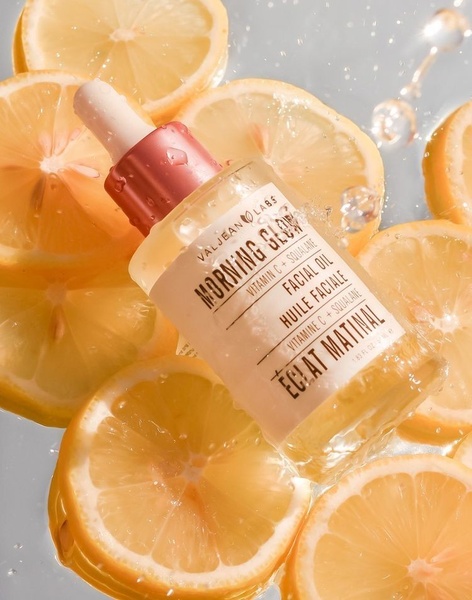 A high-quality product shot of a facial oil bottle surrounded by floating lemon slices and water droplets, creating a fresh and clean aesthetic.